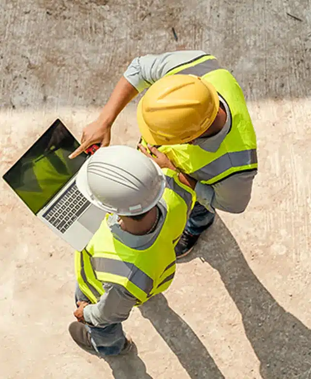 Two construction workers in hard hats and high-visibility vests reviewing project plans on laptop at job site